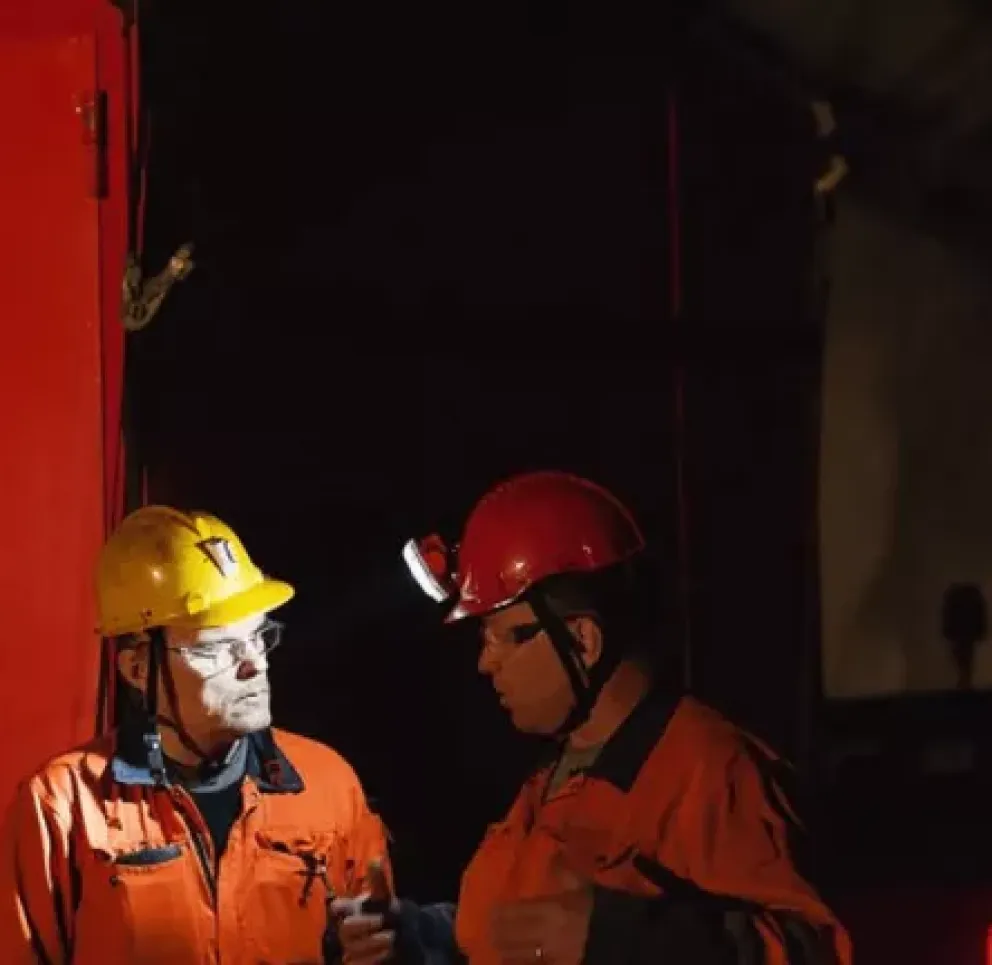 Two miners wearing orange safety gear and helmets converse in a dimly lit underground setting next to a red door.