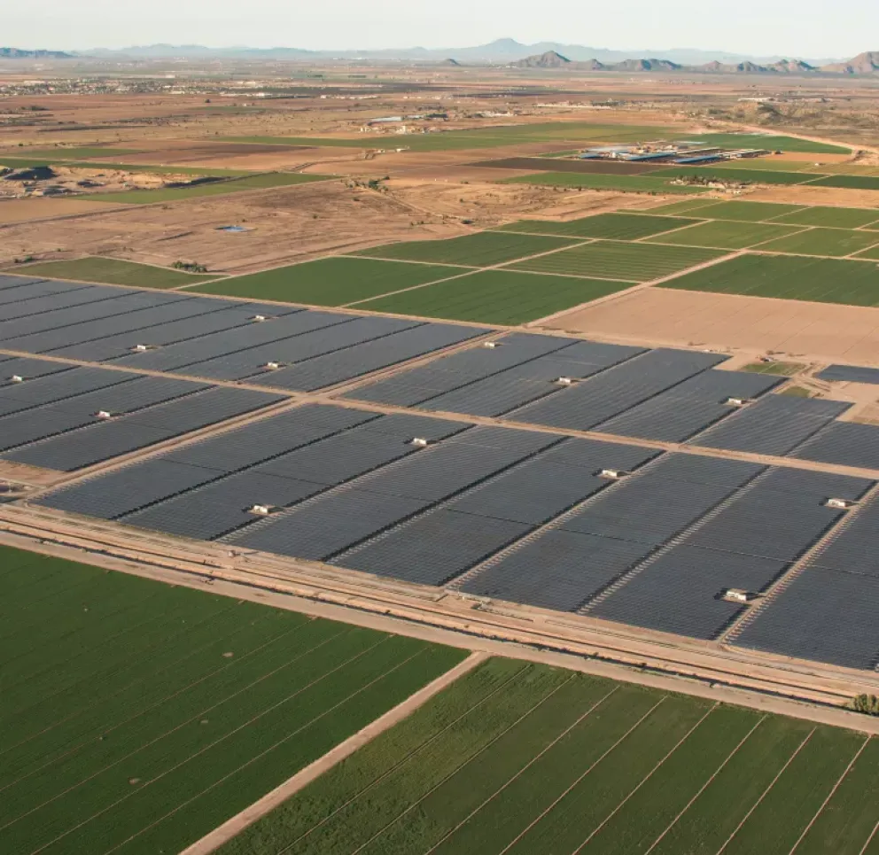 Aerial view of large solar panel arrays next to green agricultural fields in a rural landscape with distant mountains.