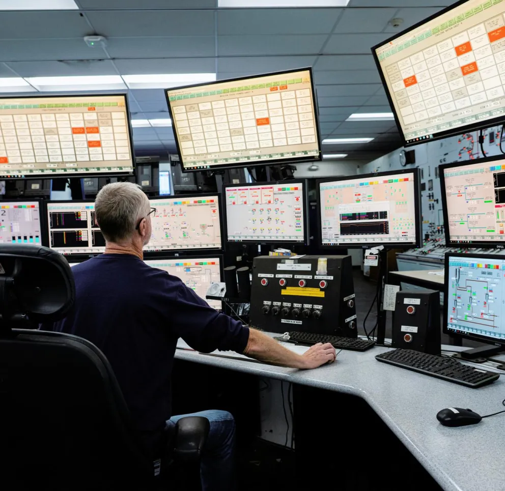 A person sits at a control room desk surrounded by multiple computer screens displaying complex data and graphs.