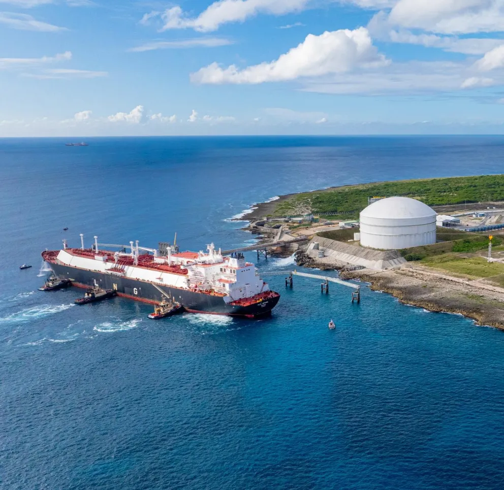 Aerial view of a large LNG tanker docked at a coastal facility with a round storage tank. The ocean is calm and extends to the horizon under a partly cloudy sky.