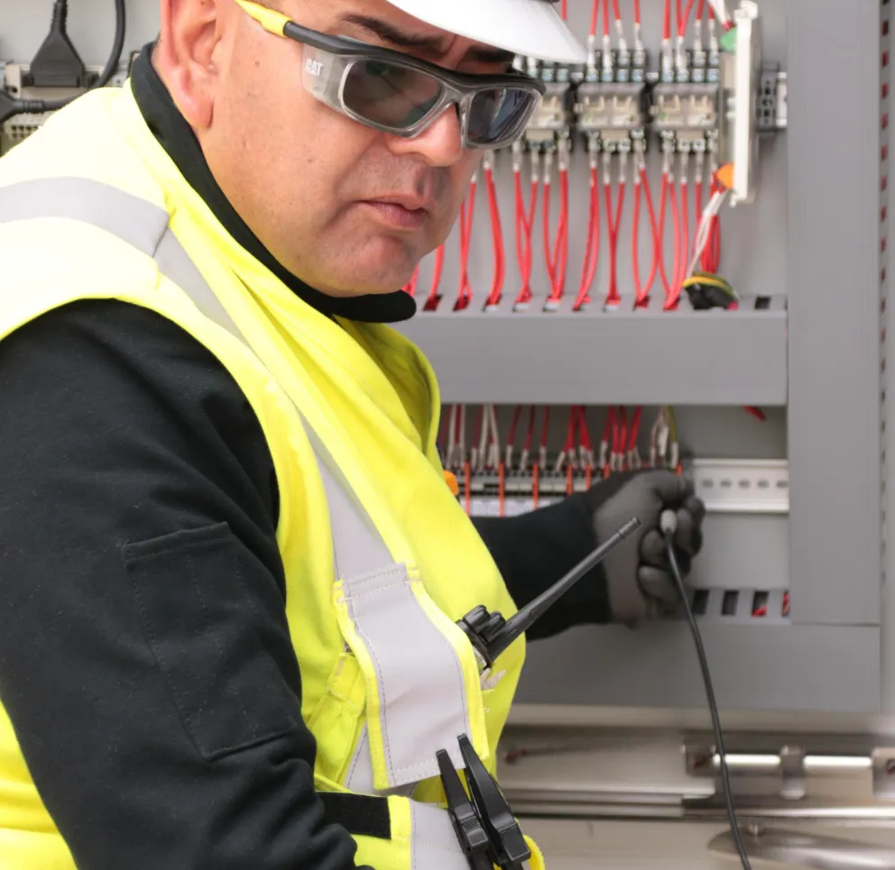 A person wearing a hard hat, safety glasses, and a high-visibility vest works on an electrical panel with red wires.