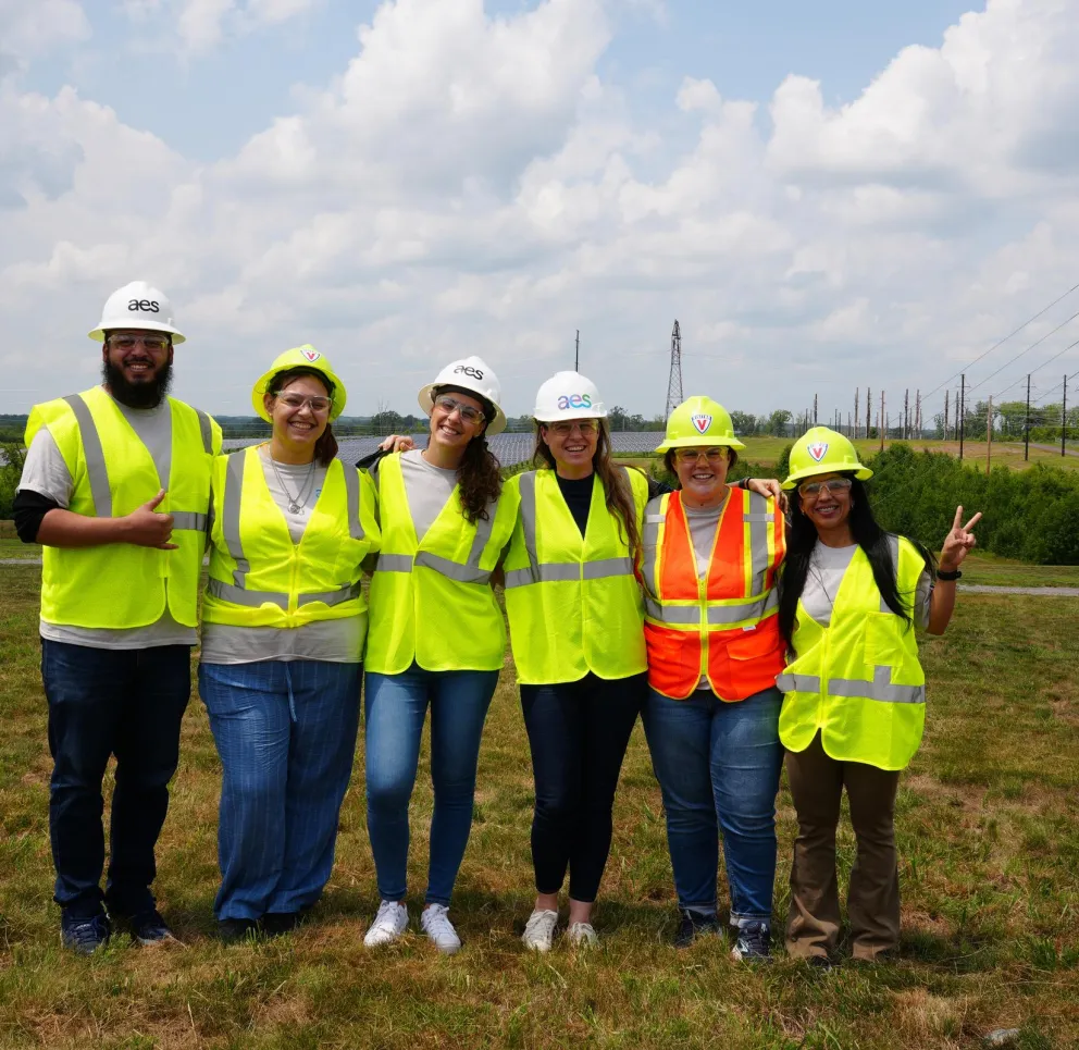 Six people wearing high-visibility vests and hard hats stand together smiling in an open grassy area with solar panels and power lines in the background. The sky is partly cloudy.