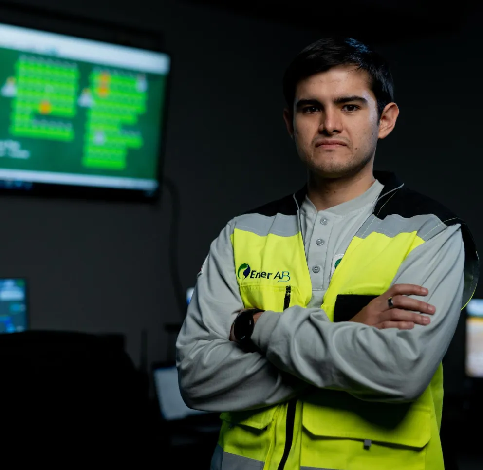 A person in a brightly colored safety vest stands with arms crossed in a control room with multiple computer screens displaying data.
