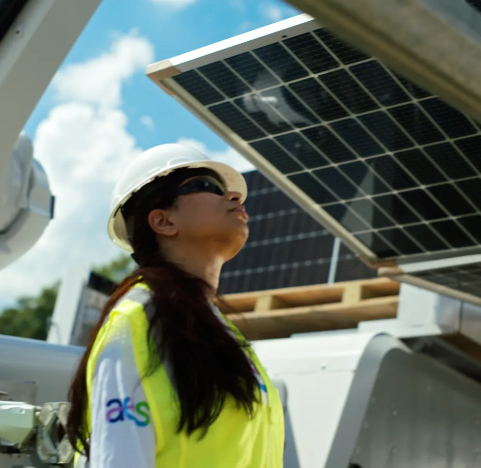 A person wearing a hard hat and safety vest inspects solar panels outdoors. The sky is clear with some clouds.