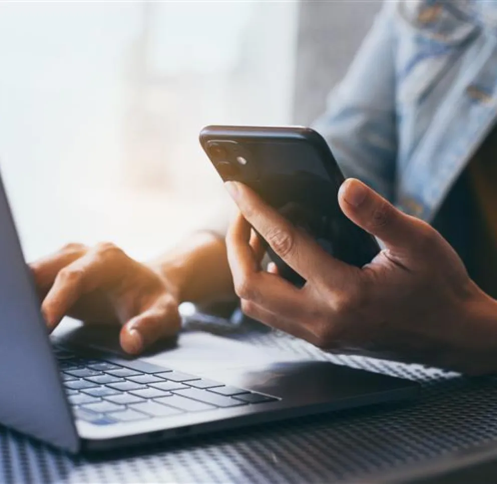 A person in a denim jacket uses a smartphone while typing on a laptop, sitting at a table with a mesh surface.
