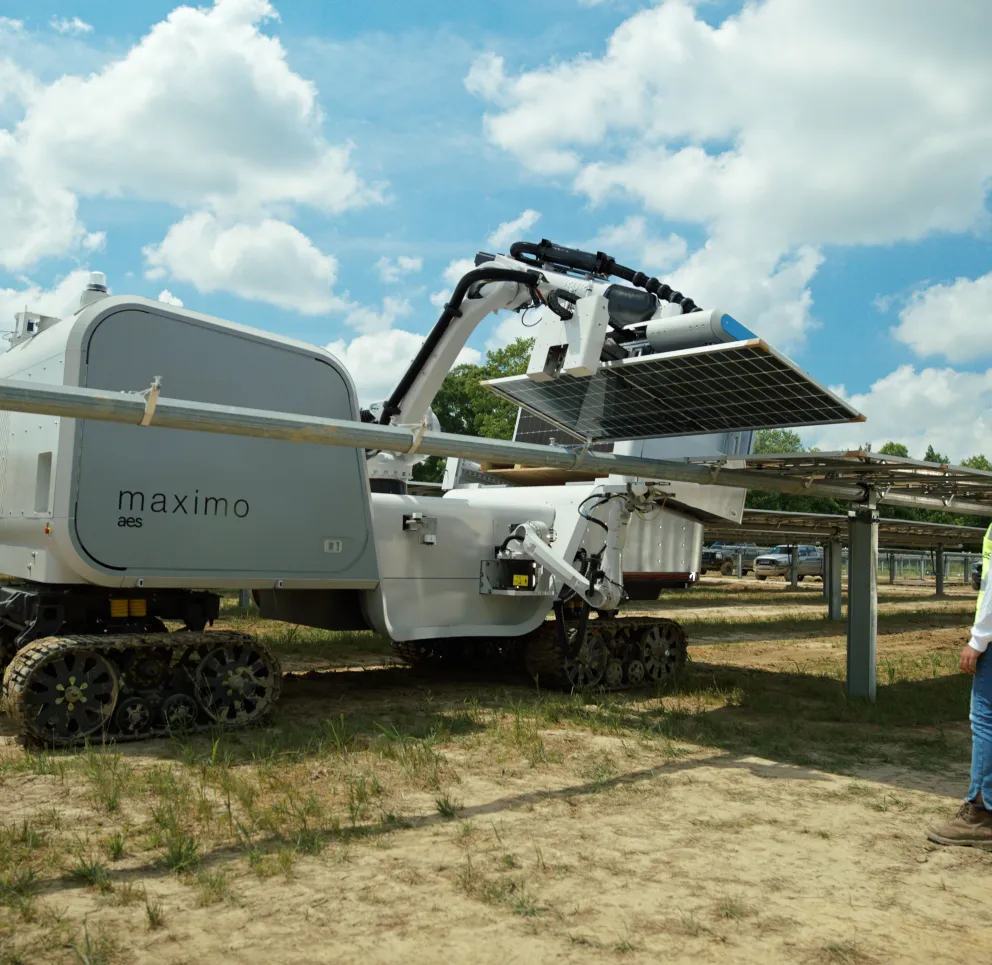 A robotic machine labeled 'maximo aes' is operating in a solar farm, adjusting a solar panel. The machine is on caterpillar tracks, and a person in a safety vest and helmet observes nearby. The background shows rows of solar panels under a partly cloudy sky.