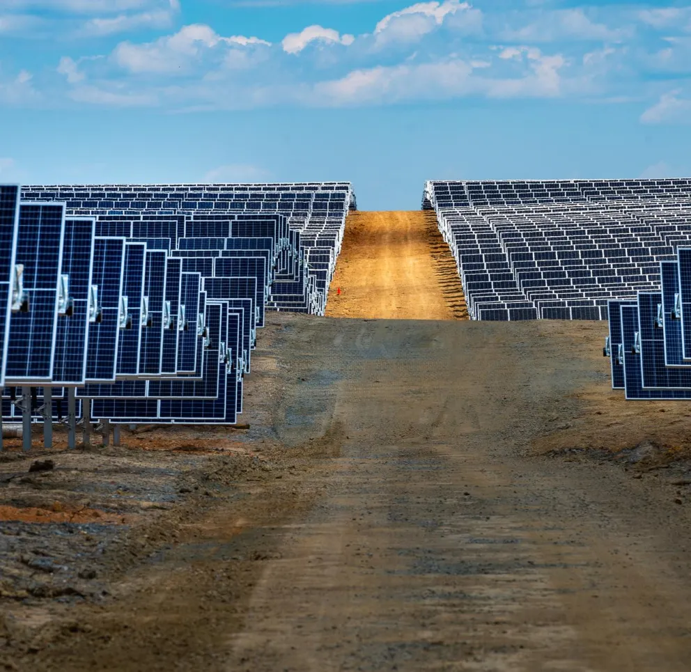 A large field of solar panels aligned in rows under a blue sky with scattered clouds, with a dirt path running through the middle.
