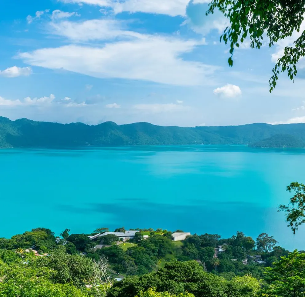 Panoramic view of a turquoise lake surrounded by lush green hills under a clear blue sky with scattered clouds.