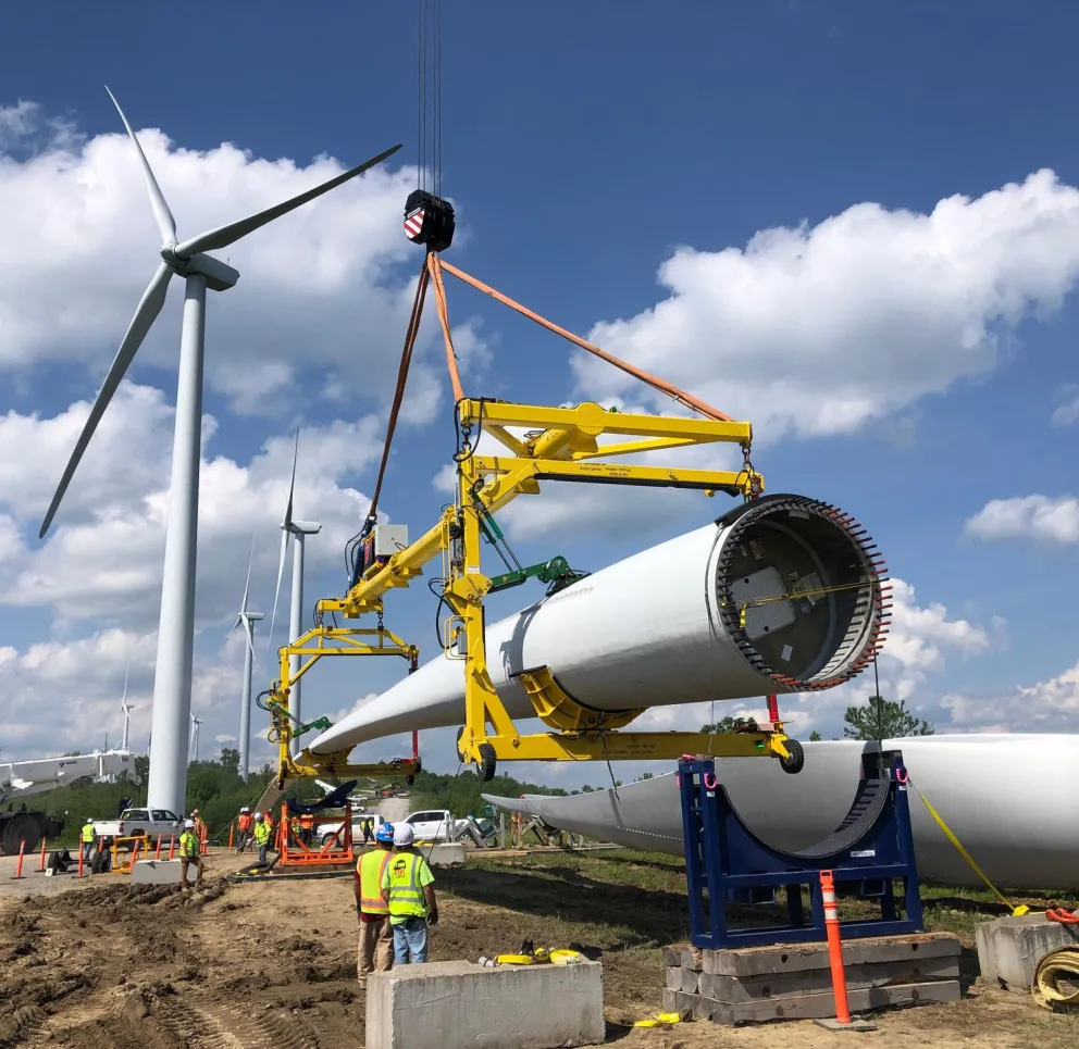 Workers in safety vests assemble a large wind turbine blade using a crane at a construction site with multiple wind turbines in the background under a blue sky with clouds.