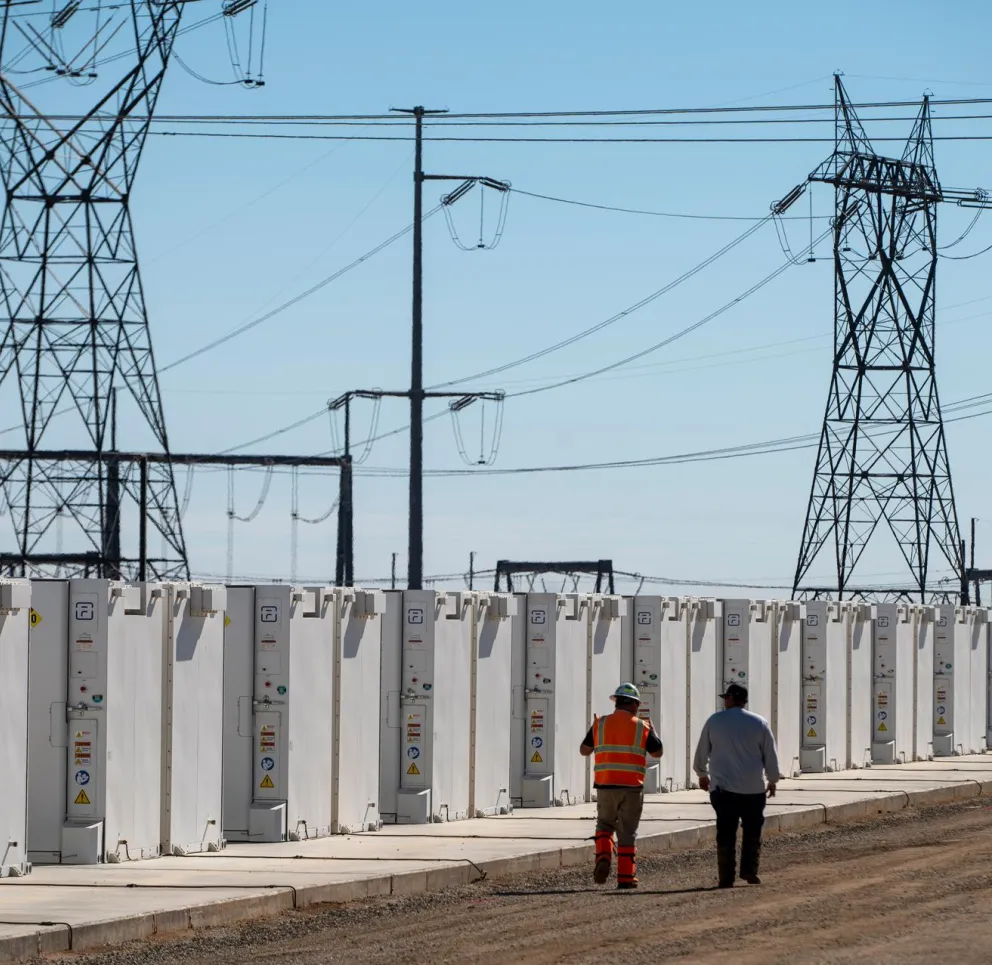 Two people walk along a row of large white battery storage units under high-voltage power lines against a clear blue sky.