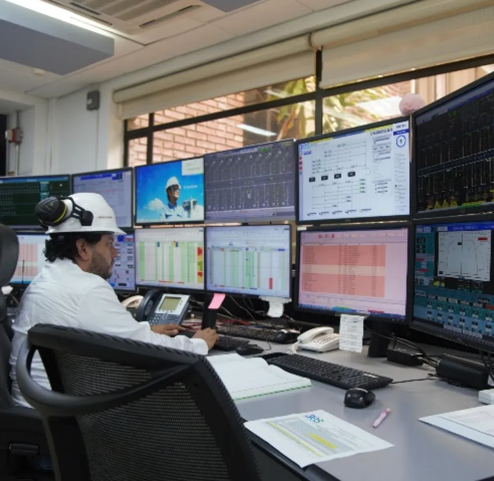 A control room with multiple monitors displaying data and graphs. A person wearing a white hard hat and headphones is seated at a desk, interacting with the computer setup. Another person in a hard hat is visible in the background. Papers and a keyboard are on the desk.