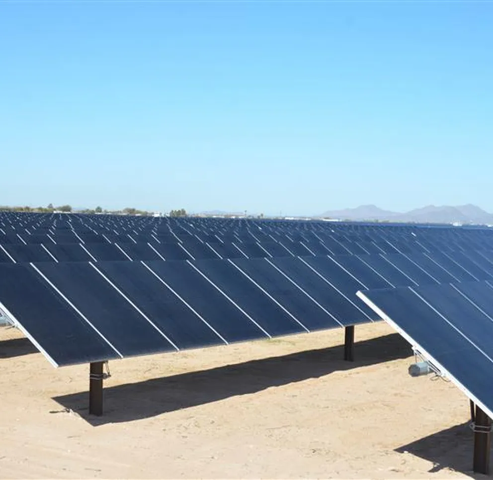 A large array of solar panels installed in a desert landscape under a clear blue sky. The panels are aligned in rows and tilted towards the sun. Mountains are visible in the distant background.