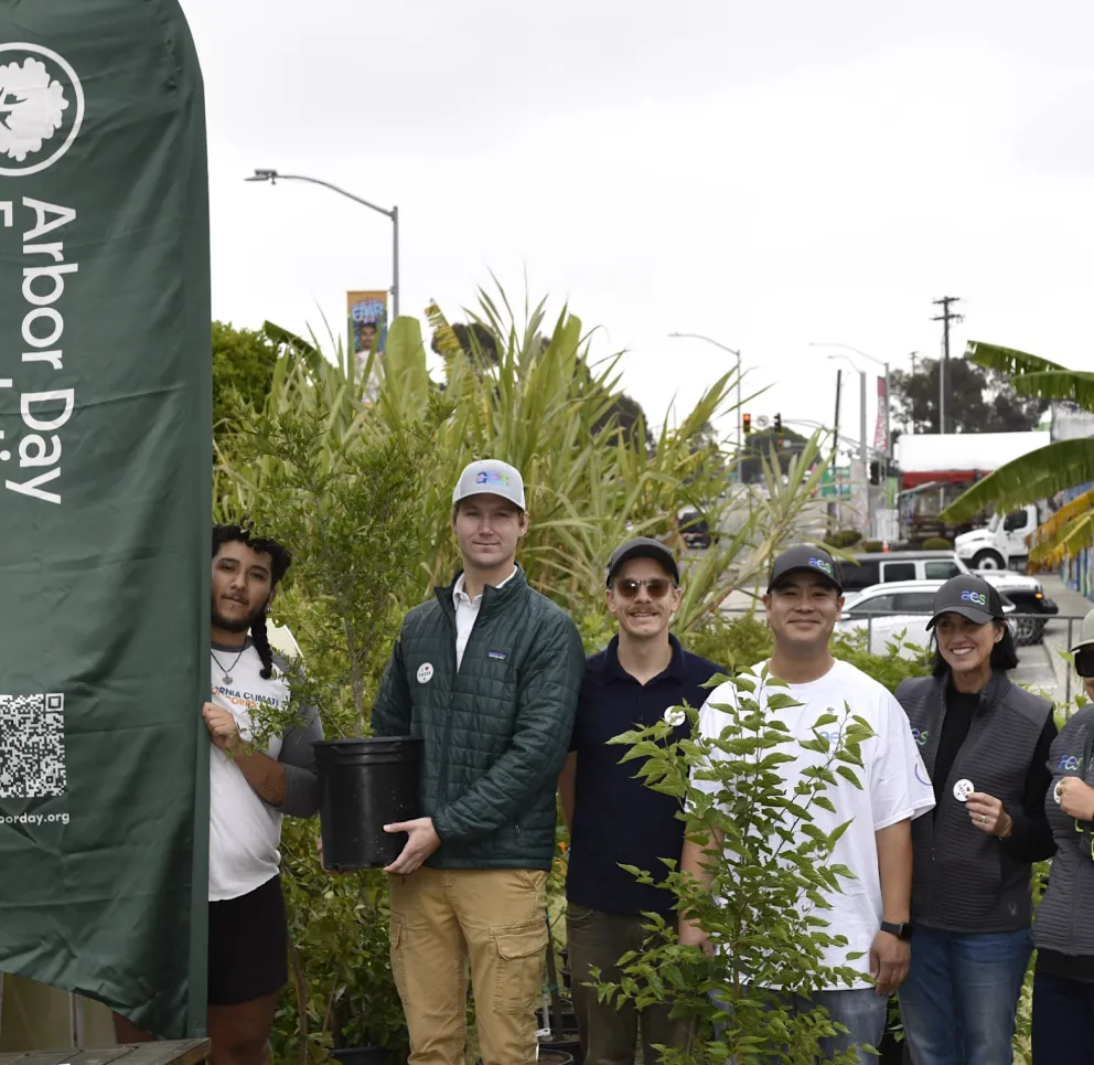 Group of people standing with plants at an Arbor Day Foundation event, smiling and holding a potted plant. A green banner with the Arbor Day Foundation logo is visible on the left.