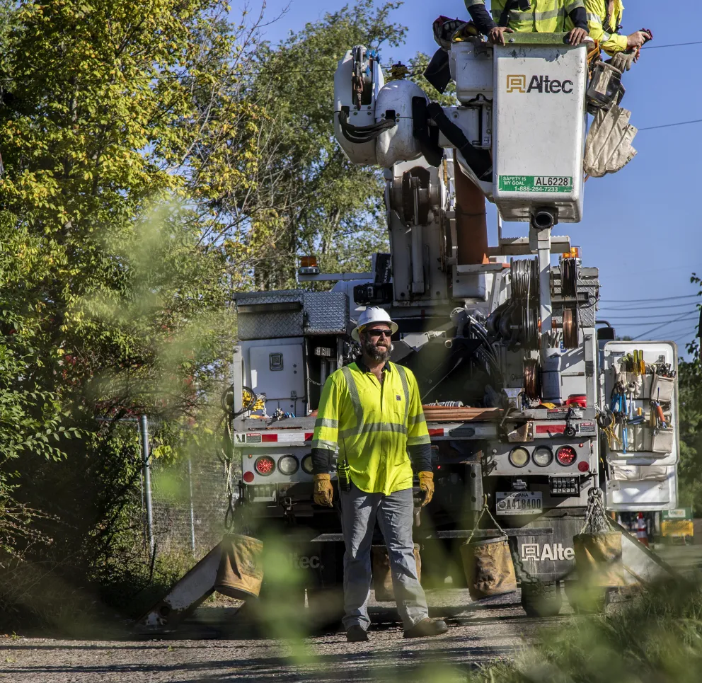 Three utility workers wearing safety gear operate a bucket truck on a rural road surrounded by trees. Two workers are in the bucket, while one stands on the ground. They are wearing high-visibility jackets and helmets, working under a clear blue sky.