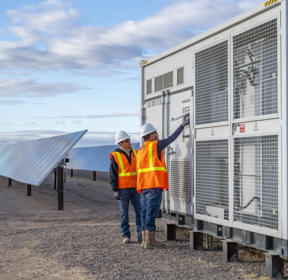 Two workers in orange safety vests and helmets operate a control panel at a solar power facility, with rows of solar panels extending into the distance under a partly cloudy sky.