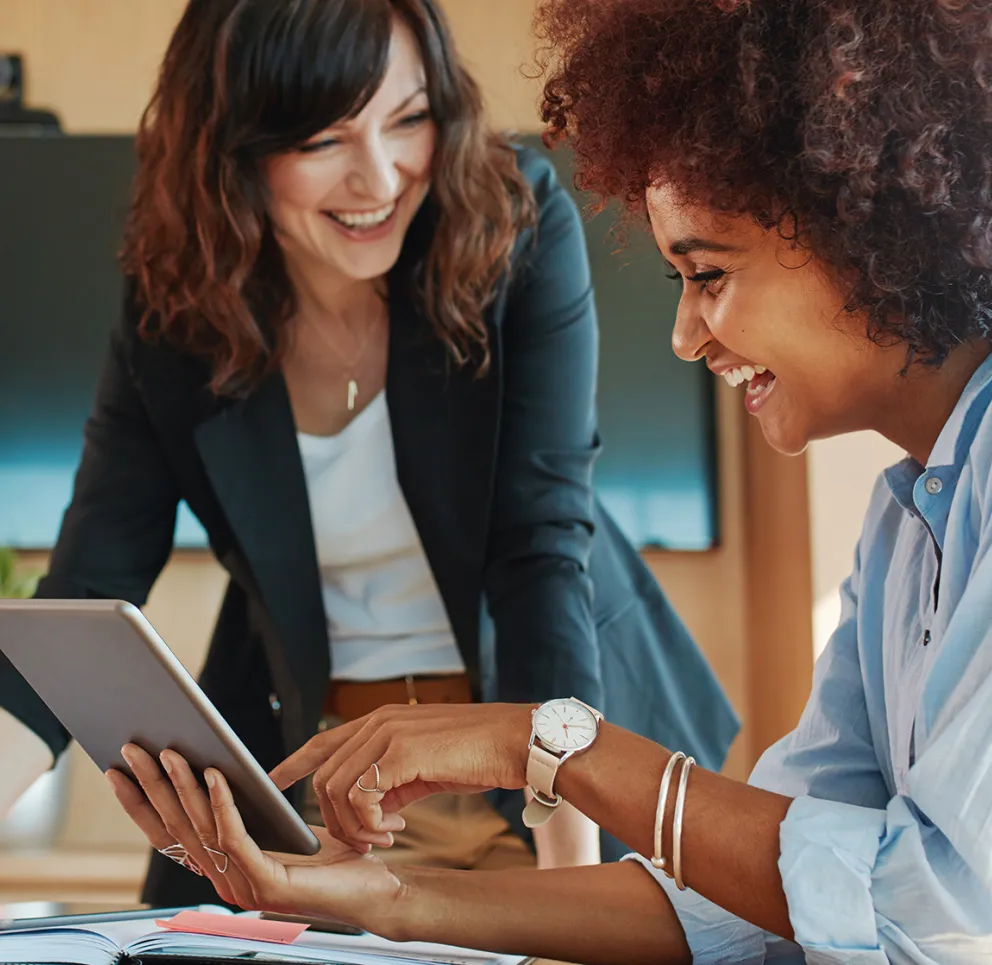 Two women smiling and collaborating in an office, one holding a tablet while the other writes notes, with a modern workspace and large screen in the background.