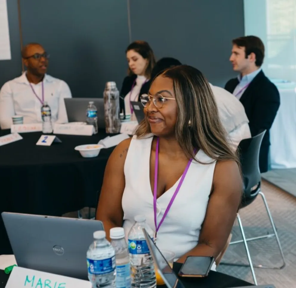 A woman wearing glasses and a white top sits at a conference table with a laptop and water bottles, smiling and listening. Other attendees are visible in the background, engaged in conversation in a modern meeting room.