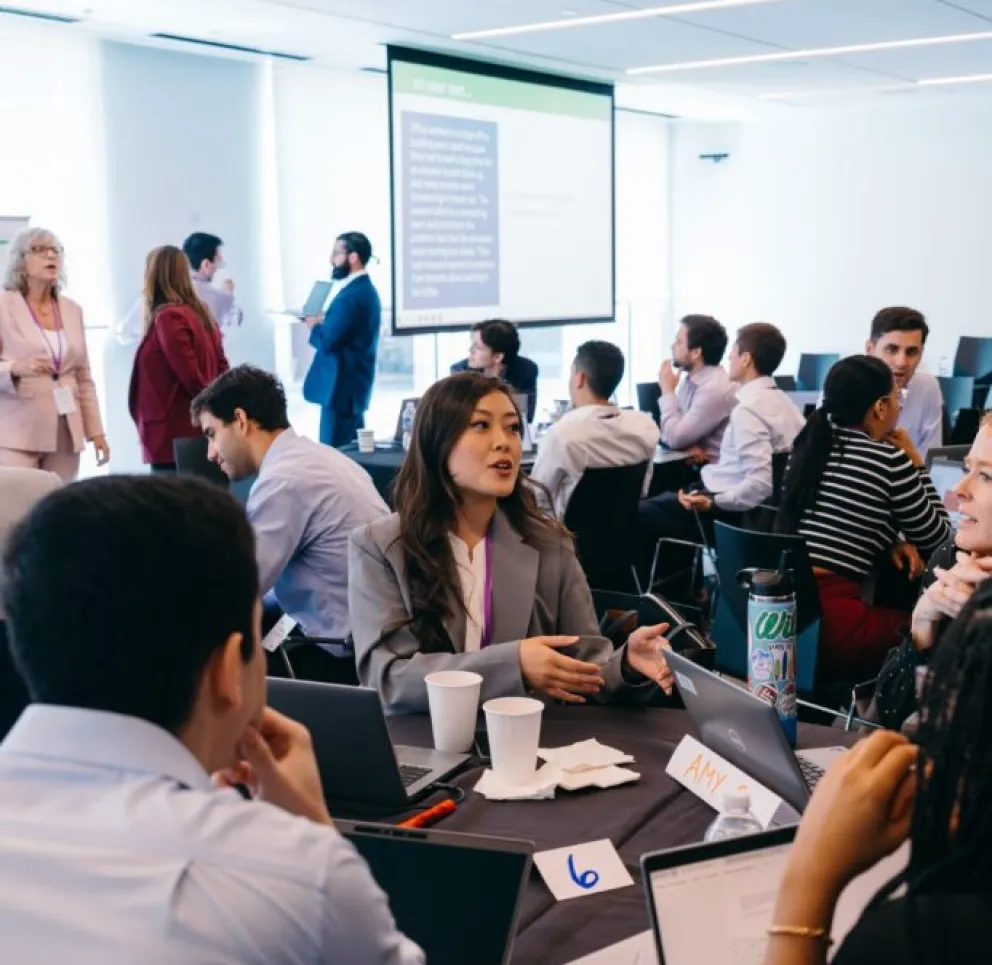 A diverse group of professionals participates in a workshop or business meeting, sitting around tables with laptops and papers, while a presentation is displayed on a screen in a modern, well-lit conference room.