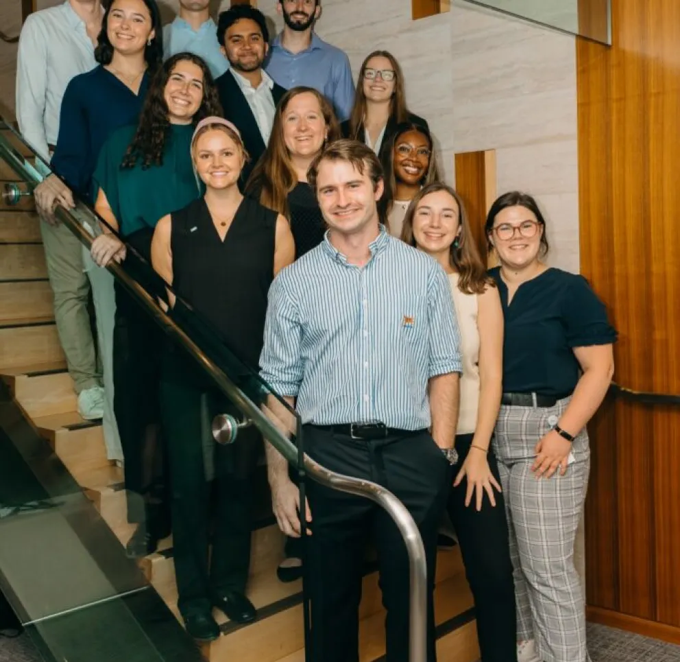 Group of thirteen young professionals smiling and posing on a modern wooden staircase with glass railing in an office setting.