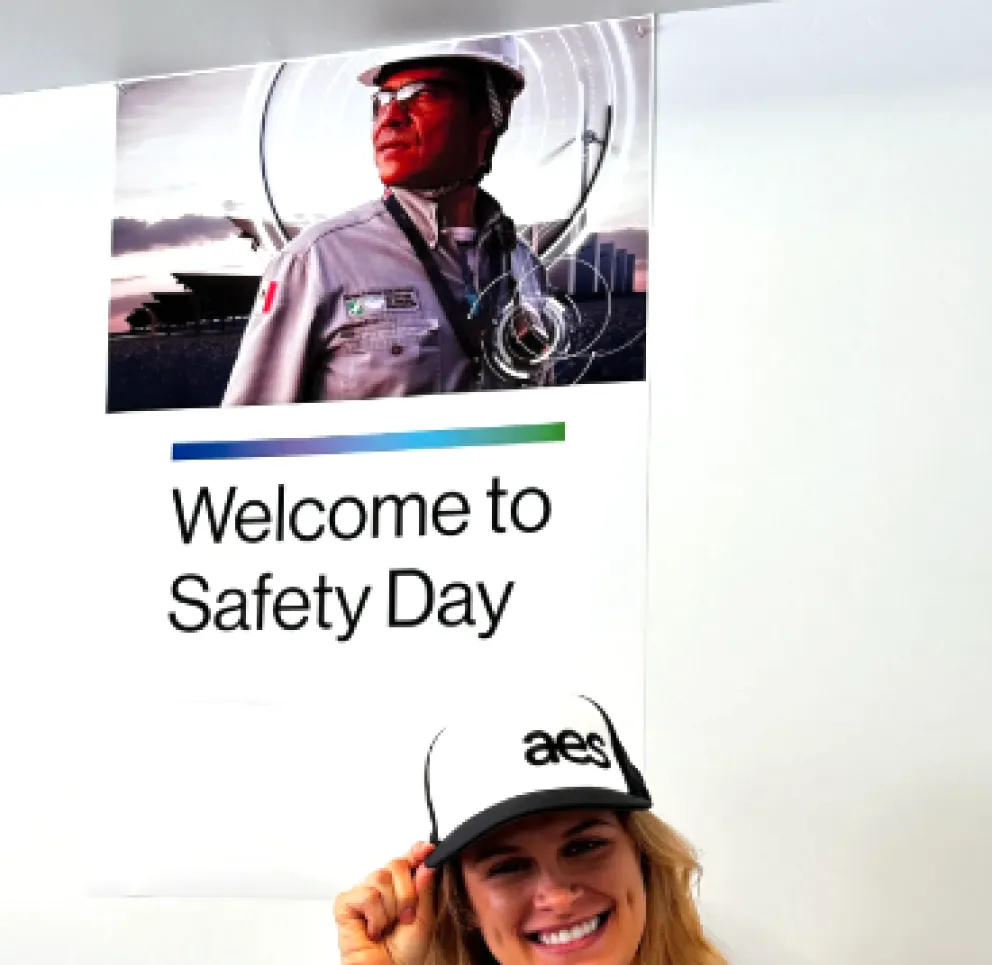 Smiling woman wearing a white AES cap stands in front of a poster that reads 'Welcome to Safety Day' with an image of a worker in safety gear.