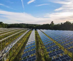 Aerial view of a large solar farm with rows of solar panels under a partly cloudy sky, surrounded by trees.