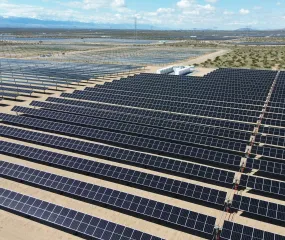 Aerial view of a large solar farm with rows of solar panels stretching across a desert landscape. Mountains are visible in the distant background under a partly cloudy sky.