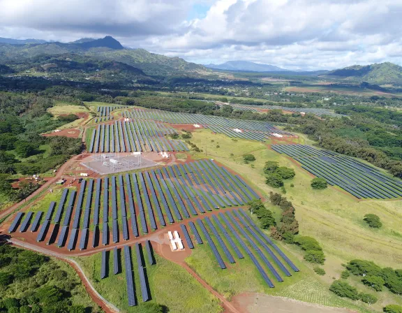 Aerial view of a large solar farm with rows of solar panels on a grassy landscape, surrounded by trees and distant mountains under a partly cloudy sky.
