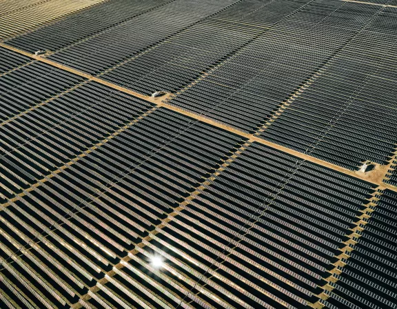 Aerial view of an expansive solar farm with rows of solar panels aligned in a grid pattern. The panels reflect sunlight, creating bright spots. Dirt paths run between the sections of panels.