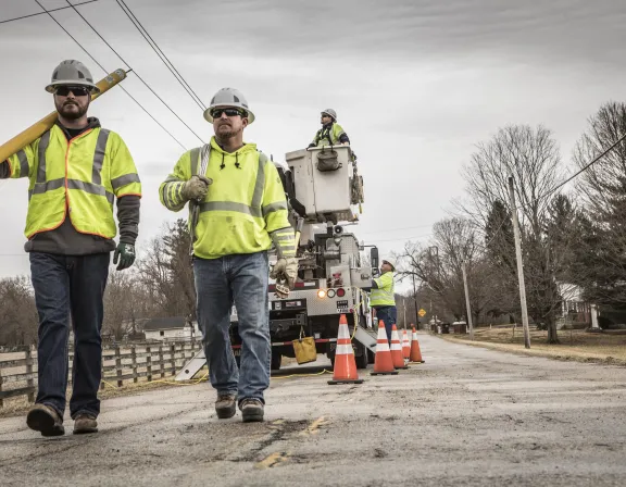 Two utility workers in high-visibility jackets and helmets walk down a rural road. One carries a large tool. In the background, a worker operates a bucket truck with safety cones placed around it. Bare trees and power lines are visible along the road.