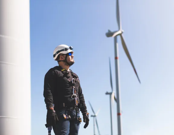 A worker wearing a safety helmet and harness stands in front of wind turbines under a clear blue sky.