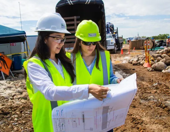 Two construction workers in high-visibility vests and hard hats examine blueprints at a construction site. The site is busy with equipment and workers in the background. One worker points to a detail on the plans while the other observes. The ground is muddy with rocks and construction cones scattered around.