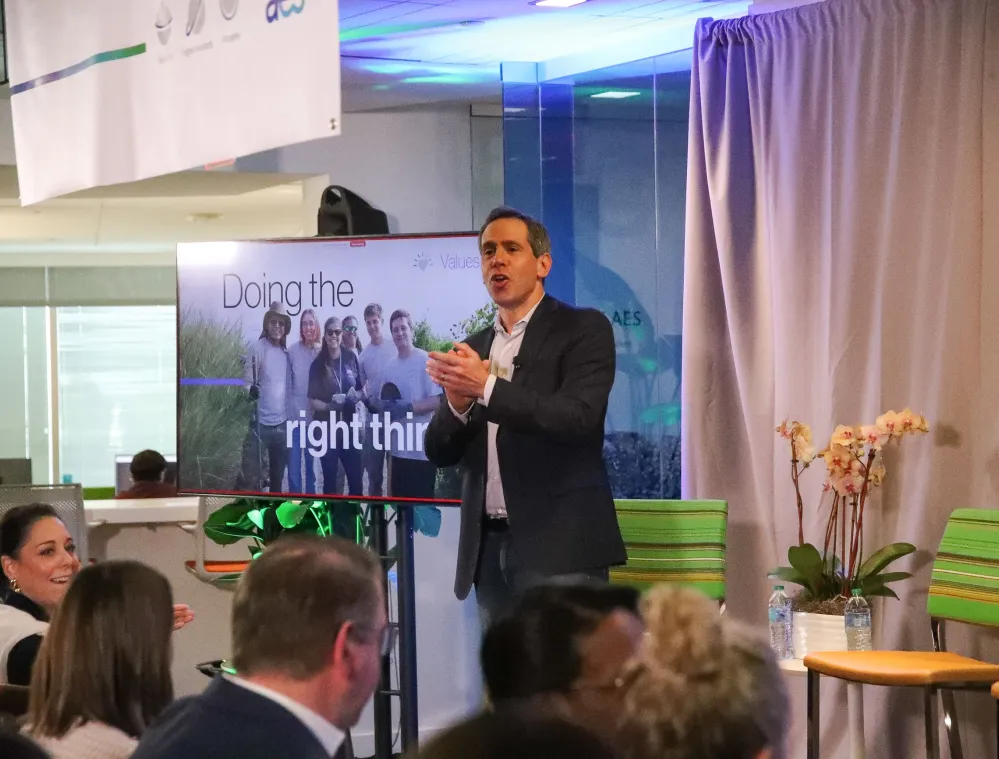 A man in a suit claps while standing next to a screen displaying the phrase 'Doing the right thing' with a group photo. People in the audience are applauding. The setting is a modern office with plants and chairs.