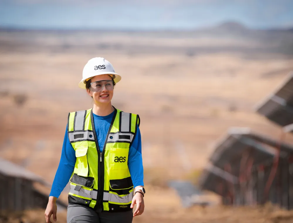 A person wearing a hard hat and safety vest smiles while walking in a solar farm with panels in the background.