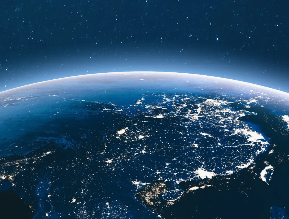 View of Earth from space at night, showing illuminated cities and networks across Asia. The curvature of the planet is visible, with a starry sky in the background.