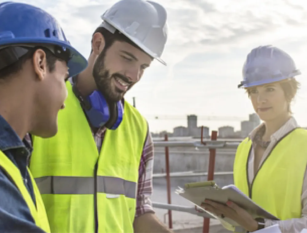 Three construction workers wearing hard hats and reflective vests are standing on a construction site. Two are engaged in conversation, while the third is holding a clipboard and looking at the camera. The background shows a city skyline and a partly cloudy sky.