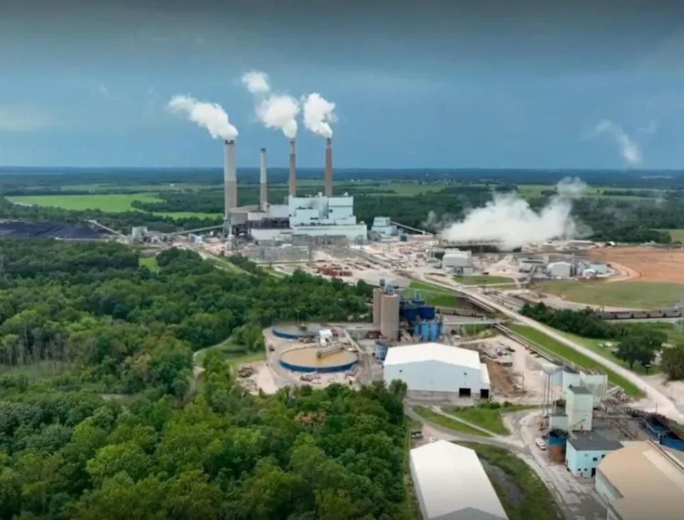 Aerial view of a large industrial power plant with multiple smokestacks emitting white smoke, surrounded by green fields and forests under a cloudy sky.