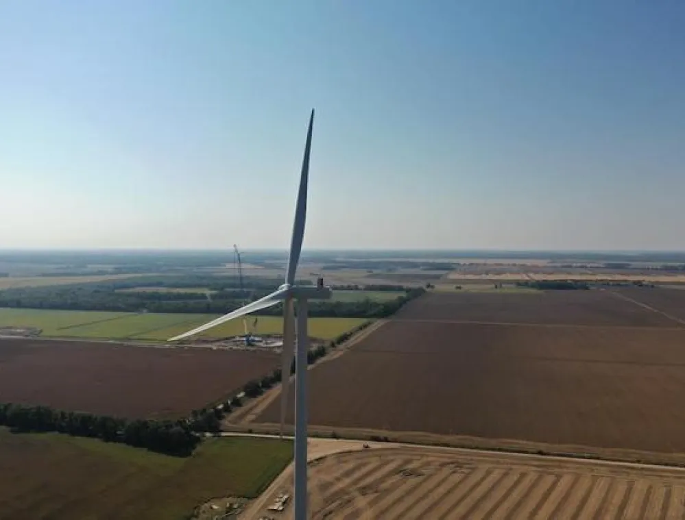 Aerial view of a wind turbine in a vast agricultural landscape with fields stretching to the horizon under a clear blue sky.