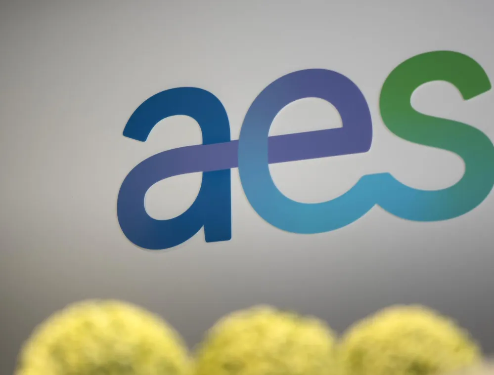 Colorful 'aes' logo on a gray background with blurred yellow-green plants in the foreground.