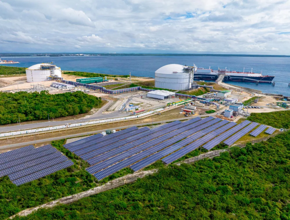 Aerial view of a coastal energy facility with large white storage tanks near a body of water. Solar panels are visible in the foreground, surrounded by greenery. Two ships are docked at the facility.