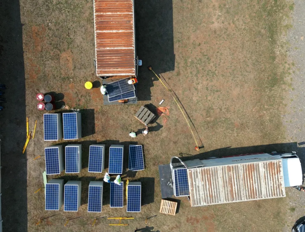 Aerial view of a solar panel installation site with several solar panels arranged on the ground, workers in hard hats, and two shipping containers. Nearby, barrels and construction materials are visible.