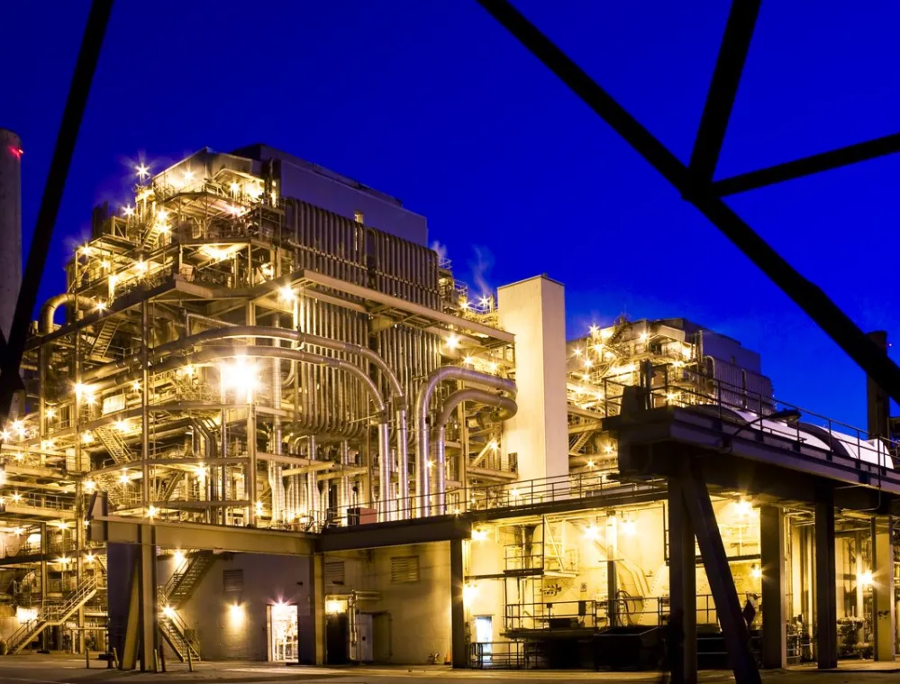 Industrial plant illuminated at night with bright lights against a deep blue sky, showcasing complex metal structures and pipes.