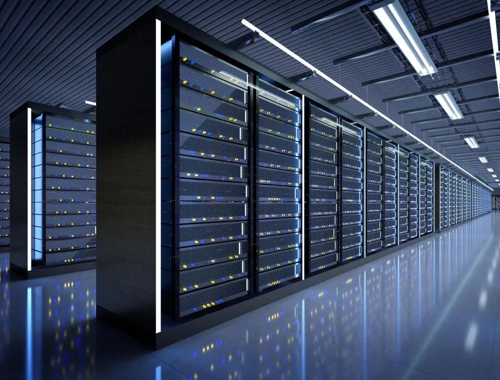 A modern data center with rows of server racks illuminated by blue and white lights. The floor is reflective, enhancing the glow from the server lights. The ceiling has evenly spaced fluorescent lights, and the room appears spacious and organized.