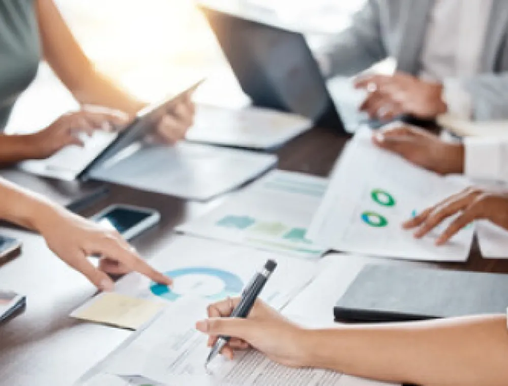 A group of people sitting around a table reviewing documents and graphs. One person points at a chart while another writes notes. Laptops and tablets are also visible on the table.