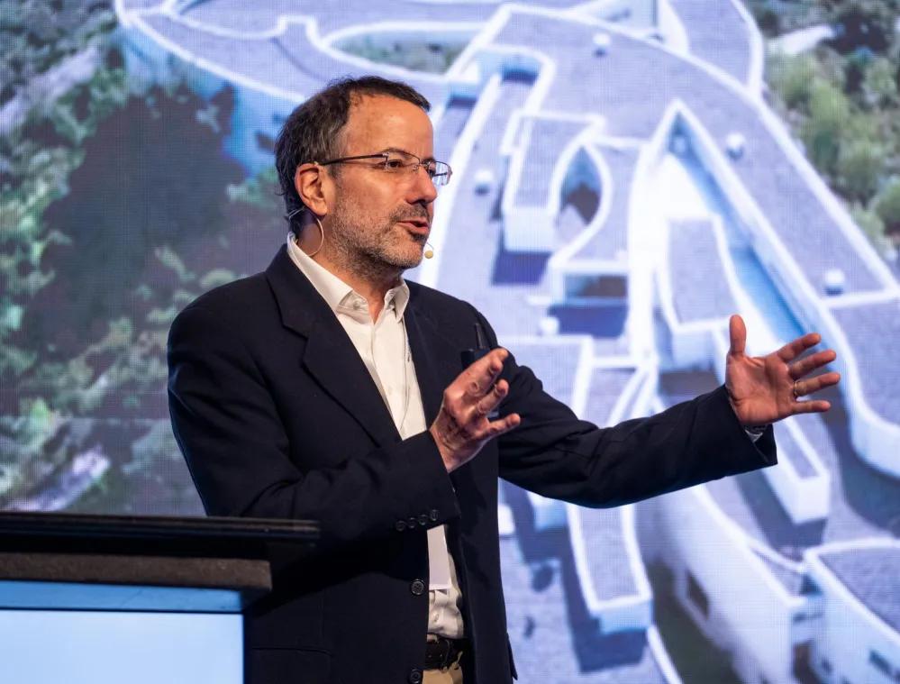 A man in a suit speaks passionately at a podium with a microphone, gesturing with his hands. A large architectural design is displayed on the screen behind him.