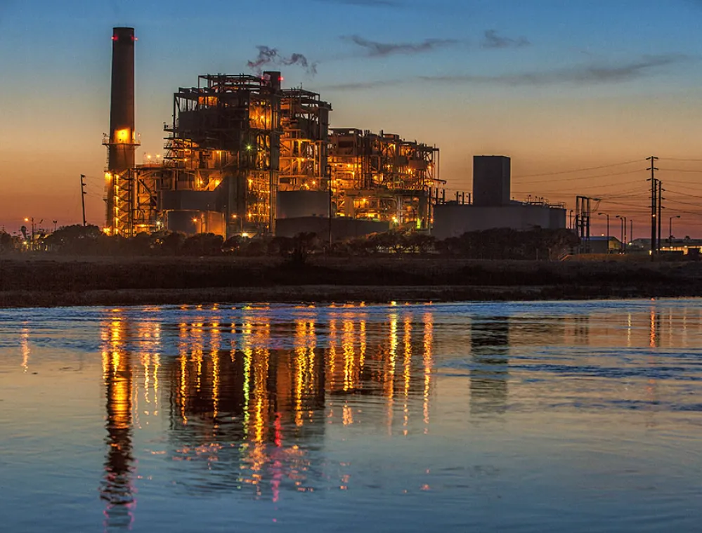 Industrial power plant illuminated at dusk, reflecting in a calm body of water.