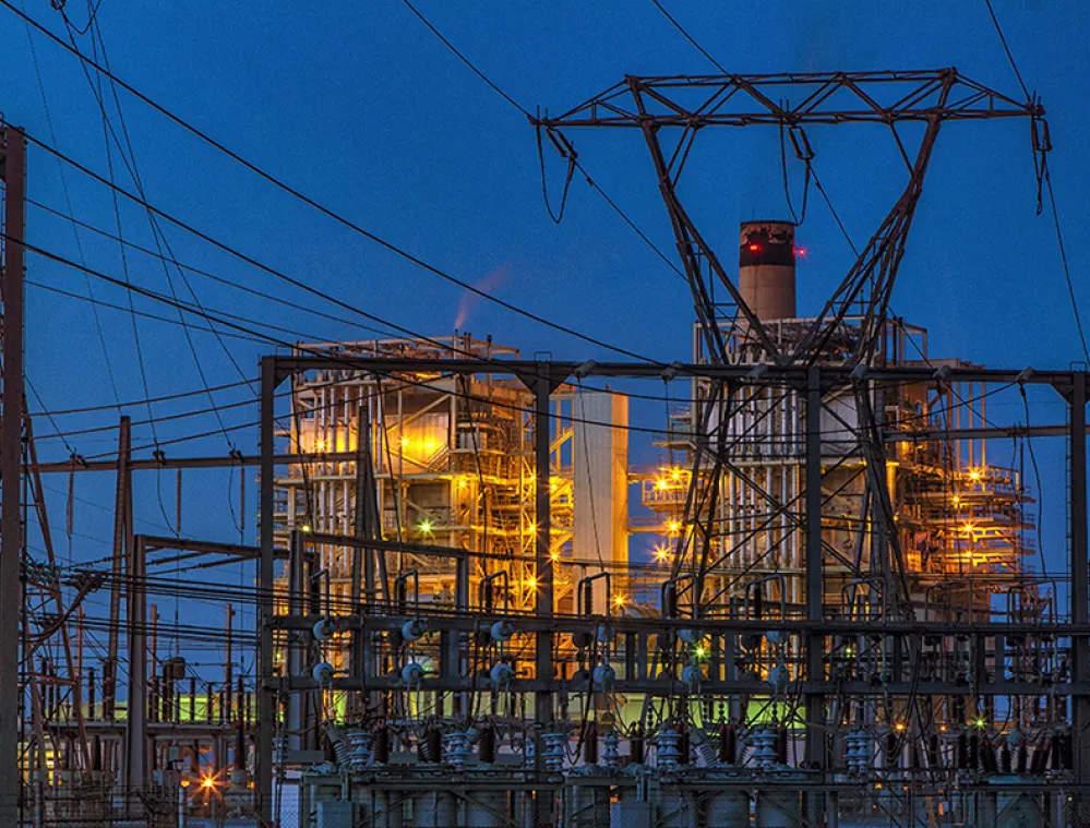 Industrial power plant at night with illuminated structures and a network of power lines against a dark blue sky.