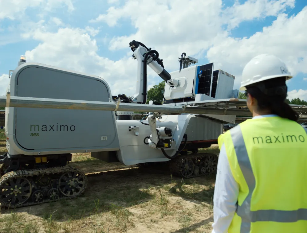 A person in a hard hat and high-visibility vest stands near a large robotic machine on tracks, labeled 'maximo aes', under a partly cloudy sky.