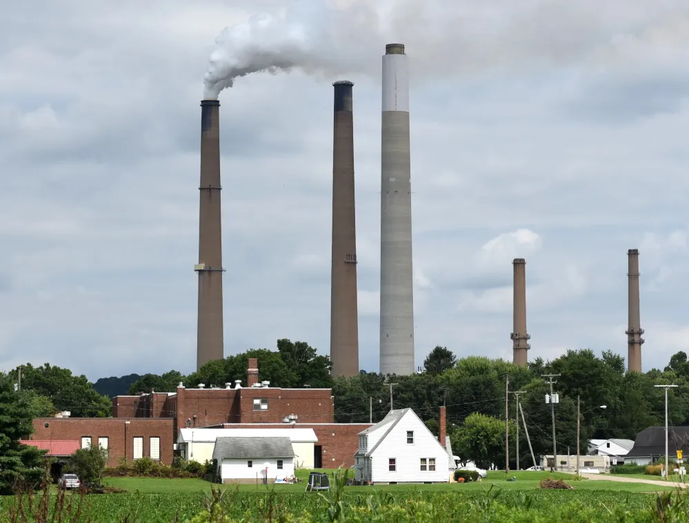 Industrial smokestacks emitting smoke against a cloudy sky, with buildings and green fields in the foreground.