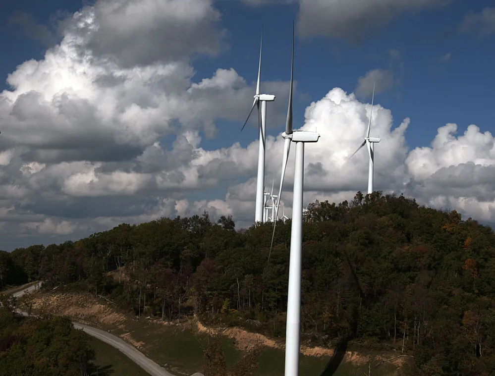 Aerial view of wind turbines on a hill surrounded by a forest under a partly cloudy sky.