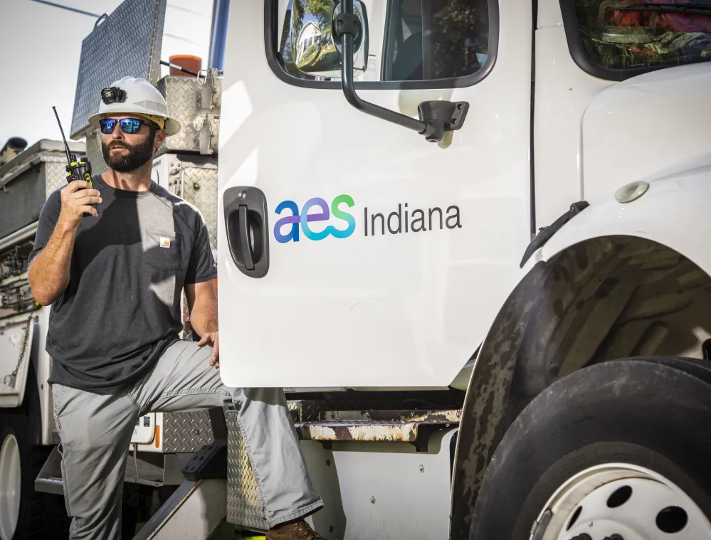 A worker wearing a helmet and sunglasses holds a radio while leaning on a utility truck with AES Indiana logo.